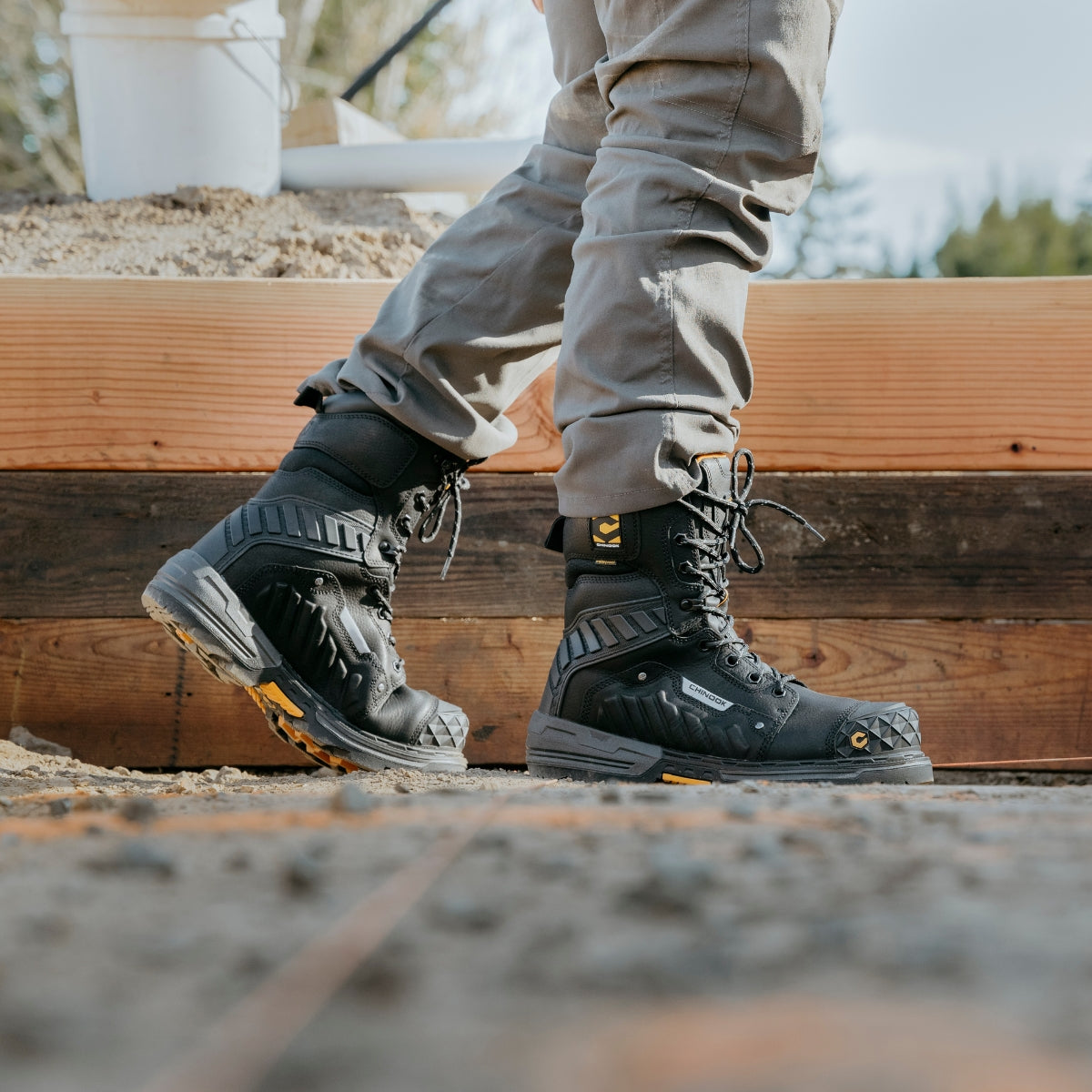 A person wearing gray pants and Chinook® Footwear Scorpion 9” Composite Toe Men’s Waterproof Work Boot - Black stands on a rough outdoor construction site with wooden beams and blurred trees in the background.