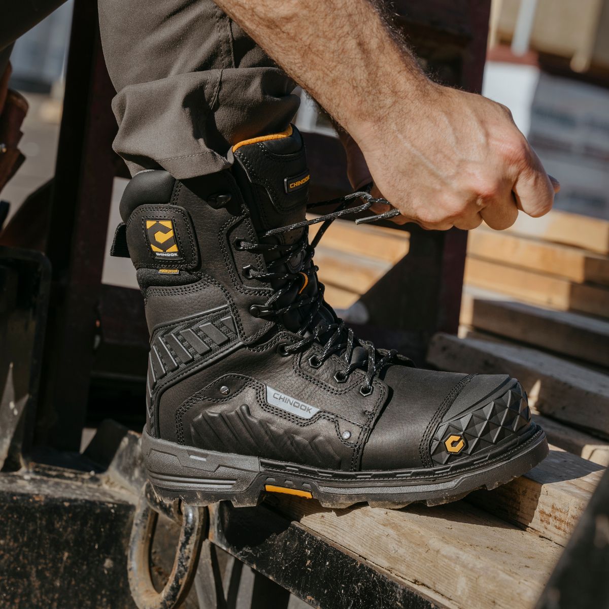 A person ties the laces of Chinook® Footwear Scorpion 9” Composite Toe Men’s Waterproof Work Boots in black, standing on industrial equipment with wooden pallets nearby. The boots feature reinforced toes and thick soles.