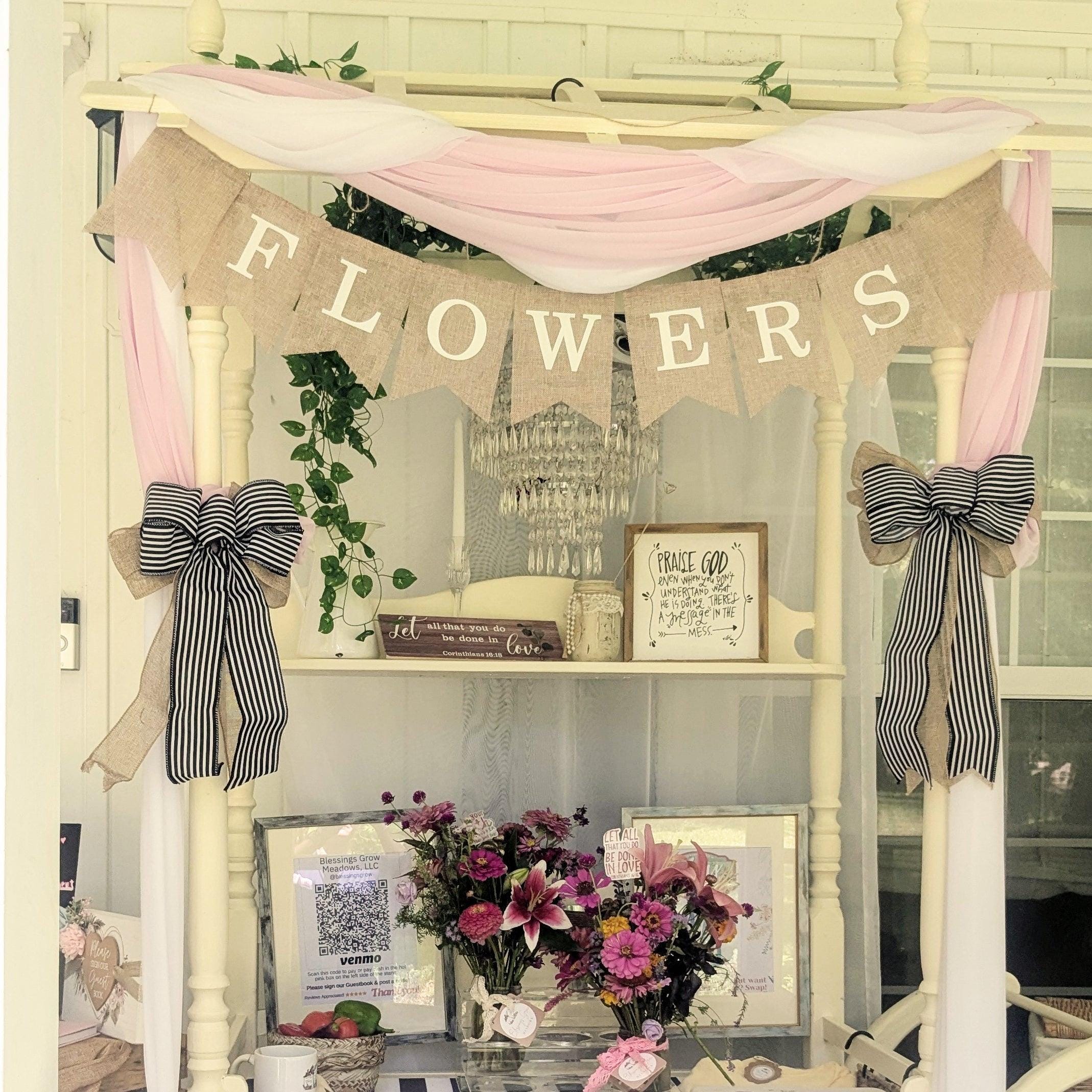 A flower shop display featuring a FLOWERS banner made of burlap, draped with pink fabric. Below are bouquets of pink and purple flowers, framed pictures, and two bows with black and white stripes. Greenery accents the arrangement.