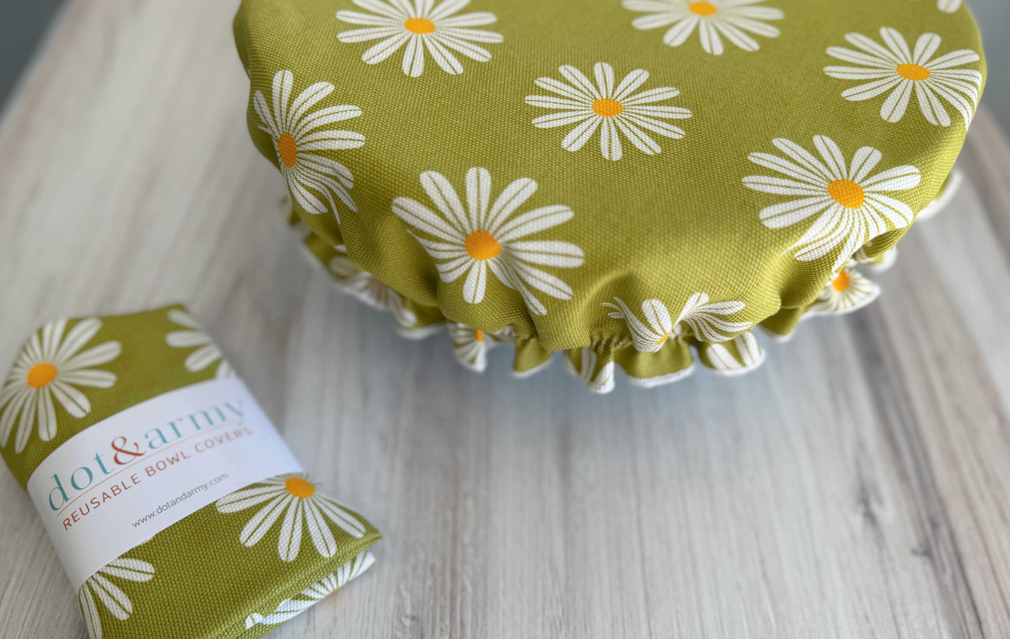 A green cotton Reusable Bowl Cover- Daisy by Dot and Army, featuring white daisies with orange centers, covers a bowl on a light wooden surface. A matching breathable cover in branded packaging sits beside it.