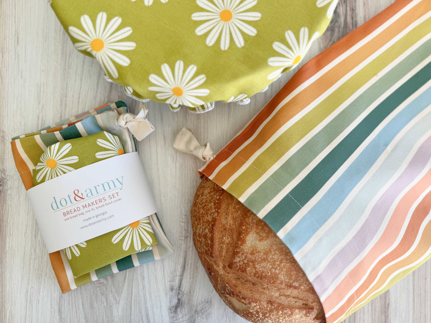A round loaf peeks from Dot and Army’s Daisy and Stripe Bread Makers Set—a linen bread bag and daisy-patterned bowl cover—displayed on a light wood surface.