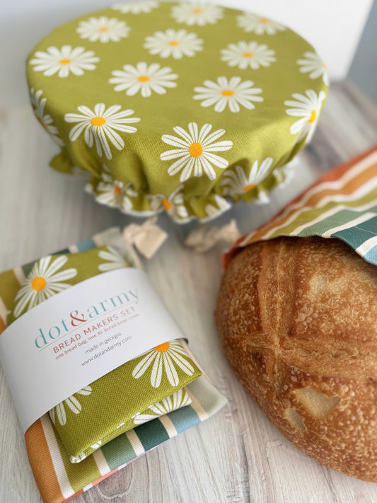A loaf of bread, a colorful striped linen blend bread bag, and a daisy-patterned bowl cover from the Dot and Army Daisy and Stripe Bread Makers Set are displayed on a light wooden surface.