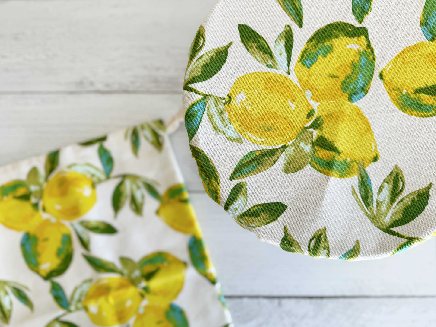 The Dot and Army Limoncello Bread Makers Set includes a bread bag and cover in white cotton, both featuring a bright yellow lemon and green leaf print—one covers a round container, while the other lies flat on a white wooden surface.