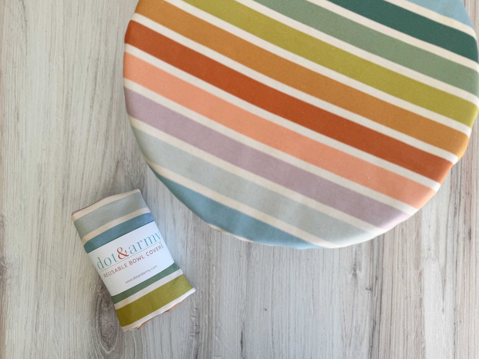 A round bowl is covered with the Dot and Army Reusable Bowl Cover in Beach Chair Stripes. Next to it, a matching bread proofing cover, rolled and labeled “dot & army,” rests on a light wood surface.