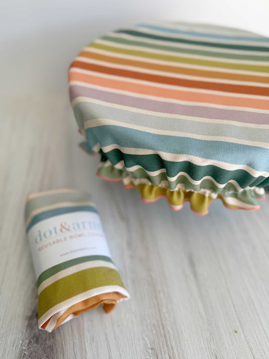 A Dot and Army Reusable Bowl Cover in Beach Chair Stripes is stretched over a bowl, with a matching folded bread proofing cover beside it on a light wooden surface.