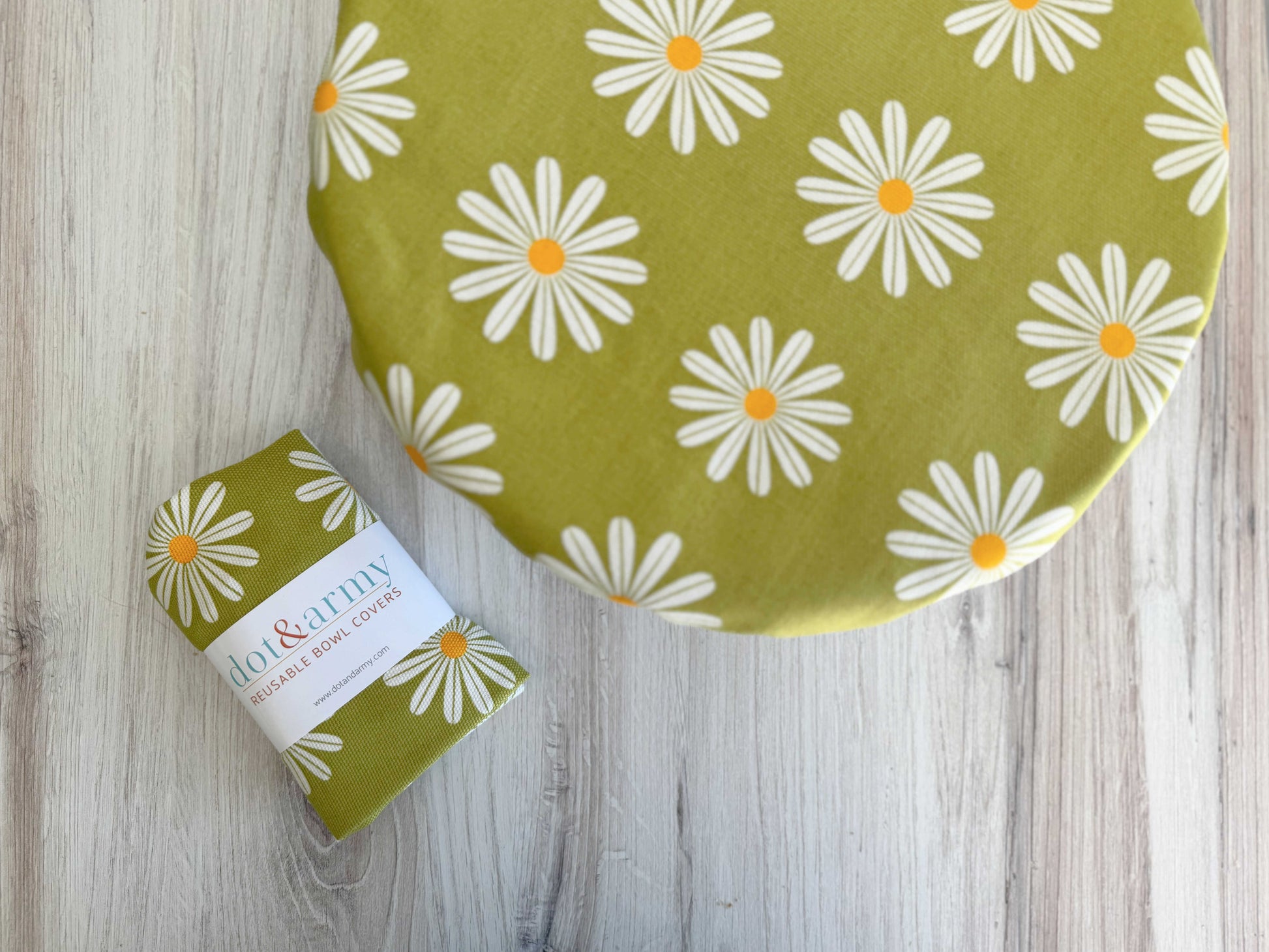 A Dot and Army Reusable Bowl Cover in a white daisy print rests on a round bowl beside a matching folded fabric, both displayed on a light wood surface.