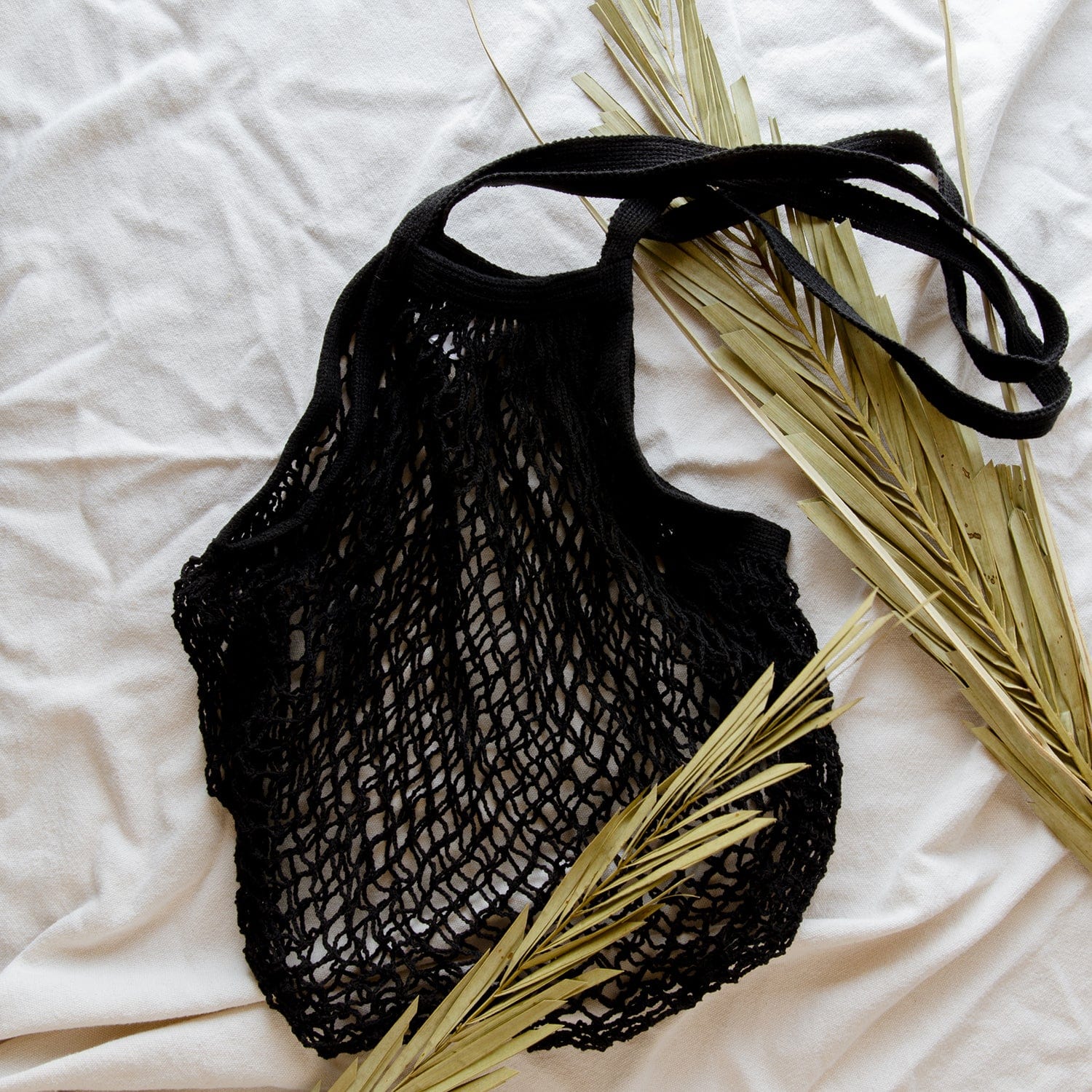 The Organic Cotton String Bag by ZeroWasteStore.com, a black zero waste mesh tote, rests on a white surface with dried palm leaves beside and partly under this reusable, plastic-free market bag.