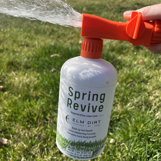 A hand sprays water with a hose attachment connected to a bottle labeled "Regenerative and Sustainable Lawn Care by Elm Dirt," applying Elm Dirt's microbe-rich organic fertilizer to green grass outdoors.