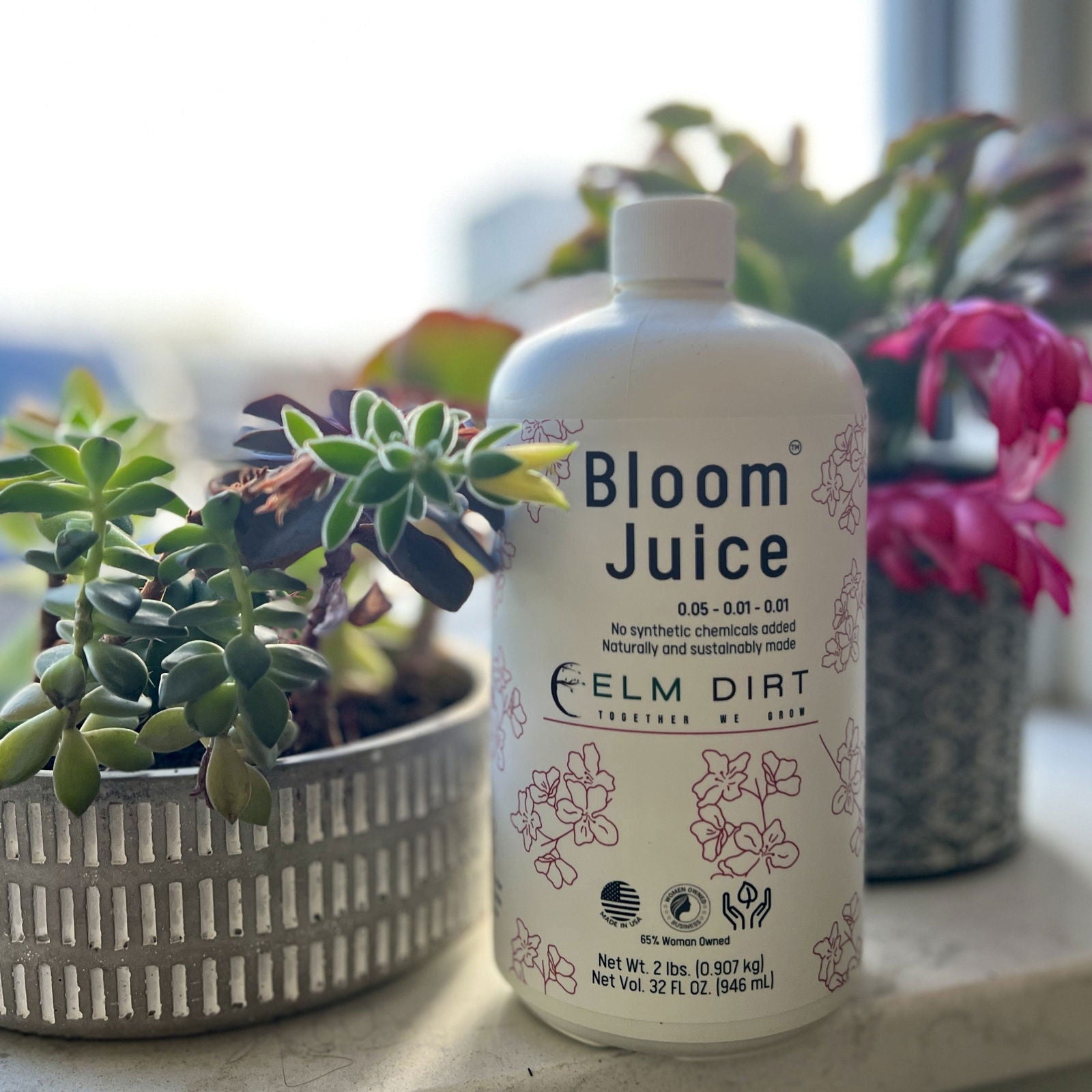 A bottle of Bloom Juice by Elm Dirt, enriched with worm castings, sits between two potted flowering plants with green and pink leaves. The white bottle features floral artwork and text, glowing in sunlight from the background.