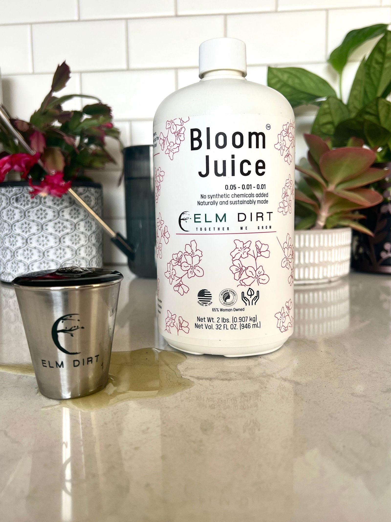 A bottle of Bloom Juice by Elm Dirt, enriched with worm castings, sits on a countertop next to an Elm Dirt measuring cup, surrounded by blooming plants and a white tile backsplash.