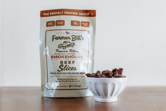 A bag of Farmer Bill's Provisions 1/2 lb Sliced Smokehouse Beef Biltong stands on a wooden surface next to a bowl of beef slices; the South Dakota packaging highlights high protein, no nitrates, and regenerative beef.