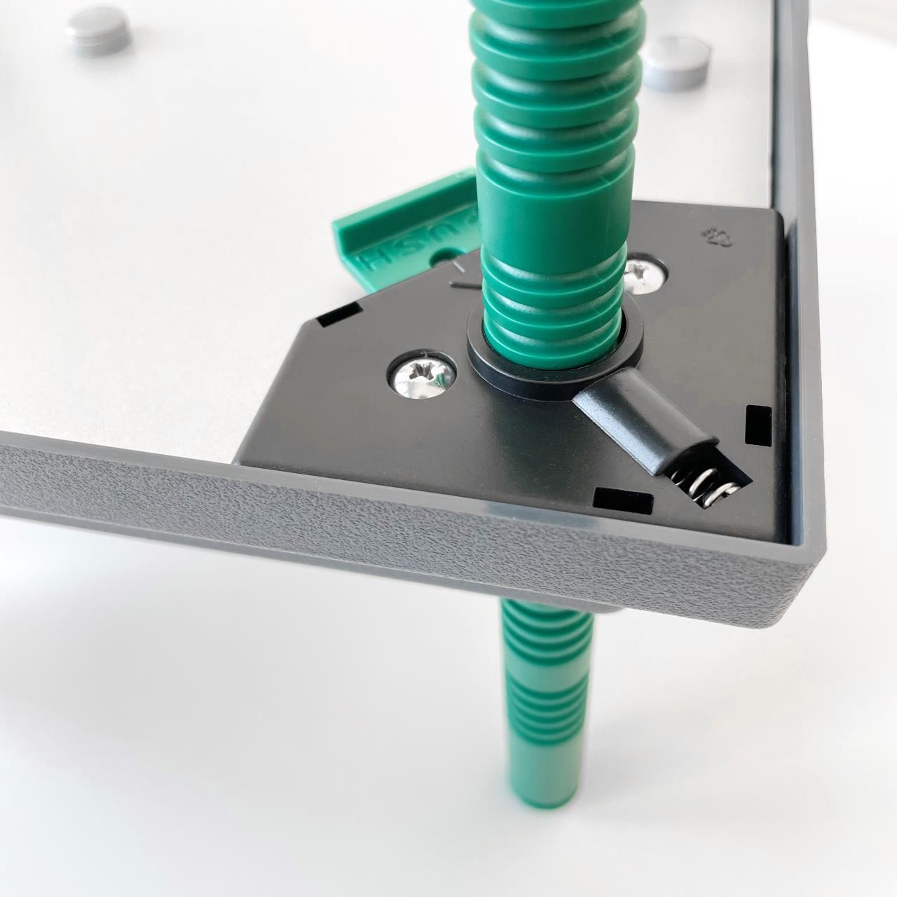 Close-up of the Farmight Chick Brooder Heater Plate by My Pet Chicken: gray panel corner with a green adjustable leg in a black bracket, secured by screws—perfect for assembling a safer chick brooder or baby chick heater.