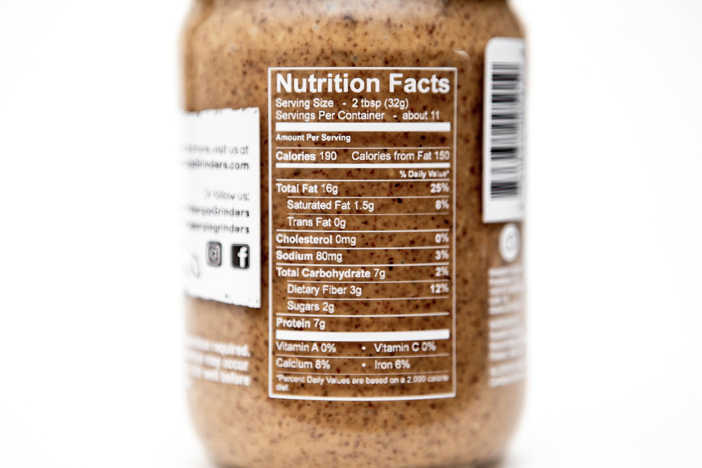 A close-up of a Georgia Grinders Honey Roasted Almond Butter jar’s nutrition label displays serving size, calories, fat, sodium, carbs, fiber, sugars, protein, and vitamins with the blurred brown almond butter visible inside.