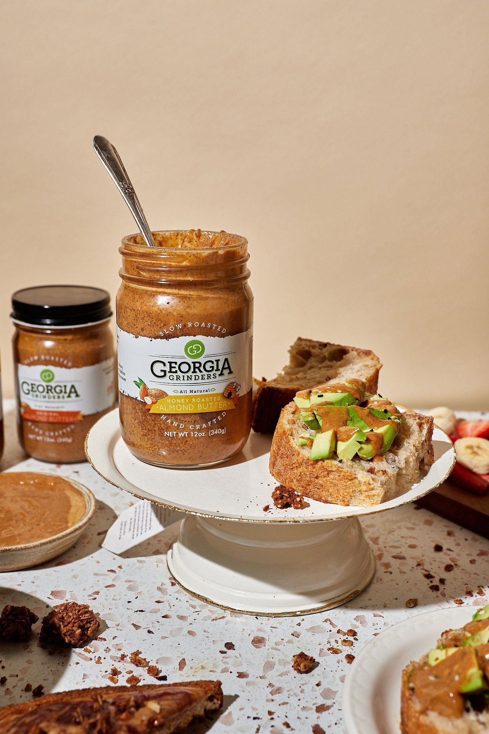 A jar from the Georgia Grinders Maple Caramel Almond Butter and Honey Roasted Almond Butter Mix Pack sits on a cake stand with a spoon inside, surrounded by bread with almond butter and apple slices; other snacks appear in the background.