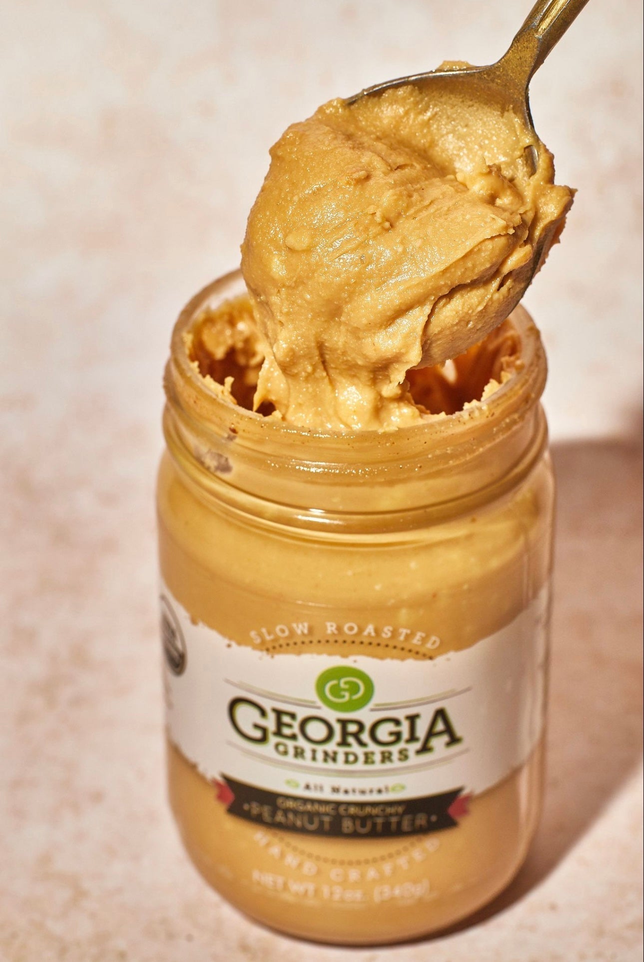 A spoon scoops chunky organic peanut butter from an open 64oz Georgia Grinders Bulk Tub of Organic Crunchy Peanut Butter (CP-CL) by Georgia Grinders, with the label shown on a light-colored surface.
