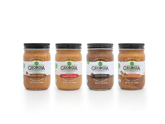 Four 12 oz jars of Georgia Grinders nut butters—Original Almond, Cashew, Pecan, and Hazelnut—stand side by side with labeled varieties and black lids. The perfect WHOLE30 Nut Butter Assortment 4 Pack (CP-CL) by Georgia Grinders.