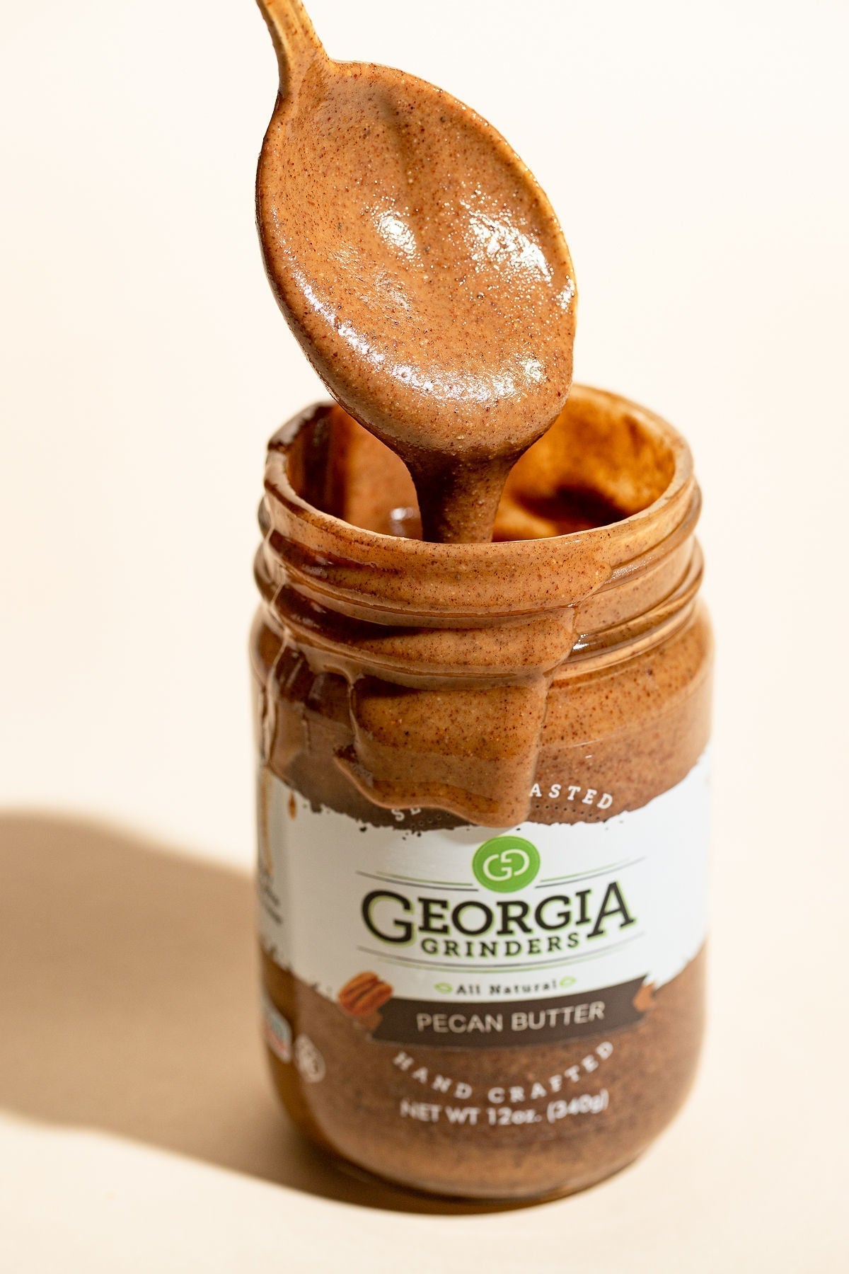 A spoon drips creamy pecan butter above an open jar from the Georgia Grinders 12 oz WHOLE30 Nut Butter Assortment 4 Pack, featuring Original Almond, Cashew, Pecan, and Hazelnut Butters.