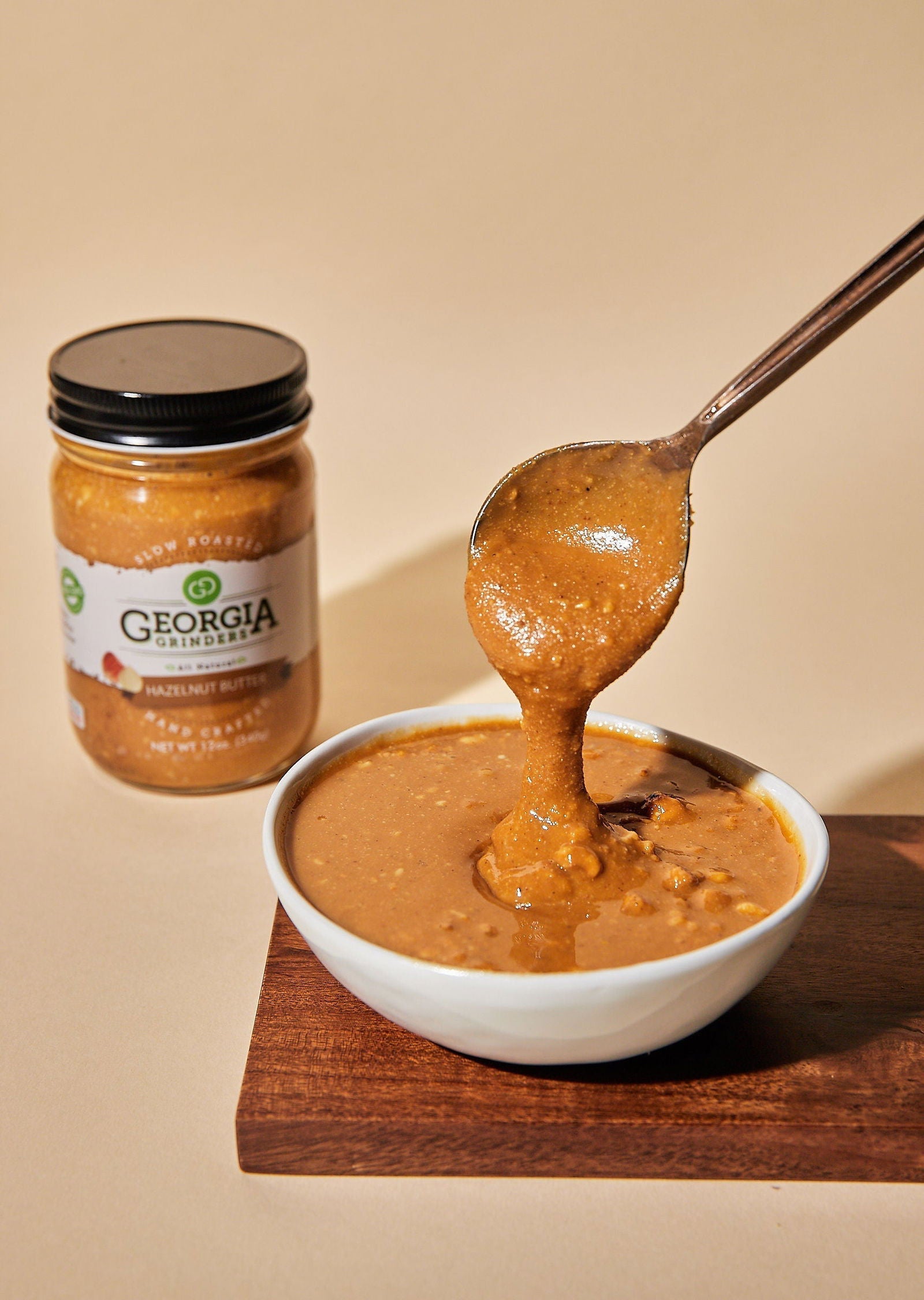 A spoon lifts thick WHOLE30 Nut Butter from a bowl on a wooden board. In the background, the Georgia Grinders 12oz WHOLE30 Nut Butter Assortment 4-Pack jar is visible against a beige backdrop.