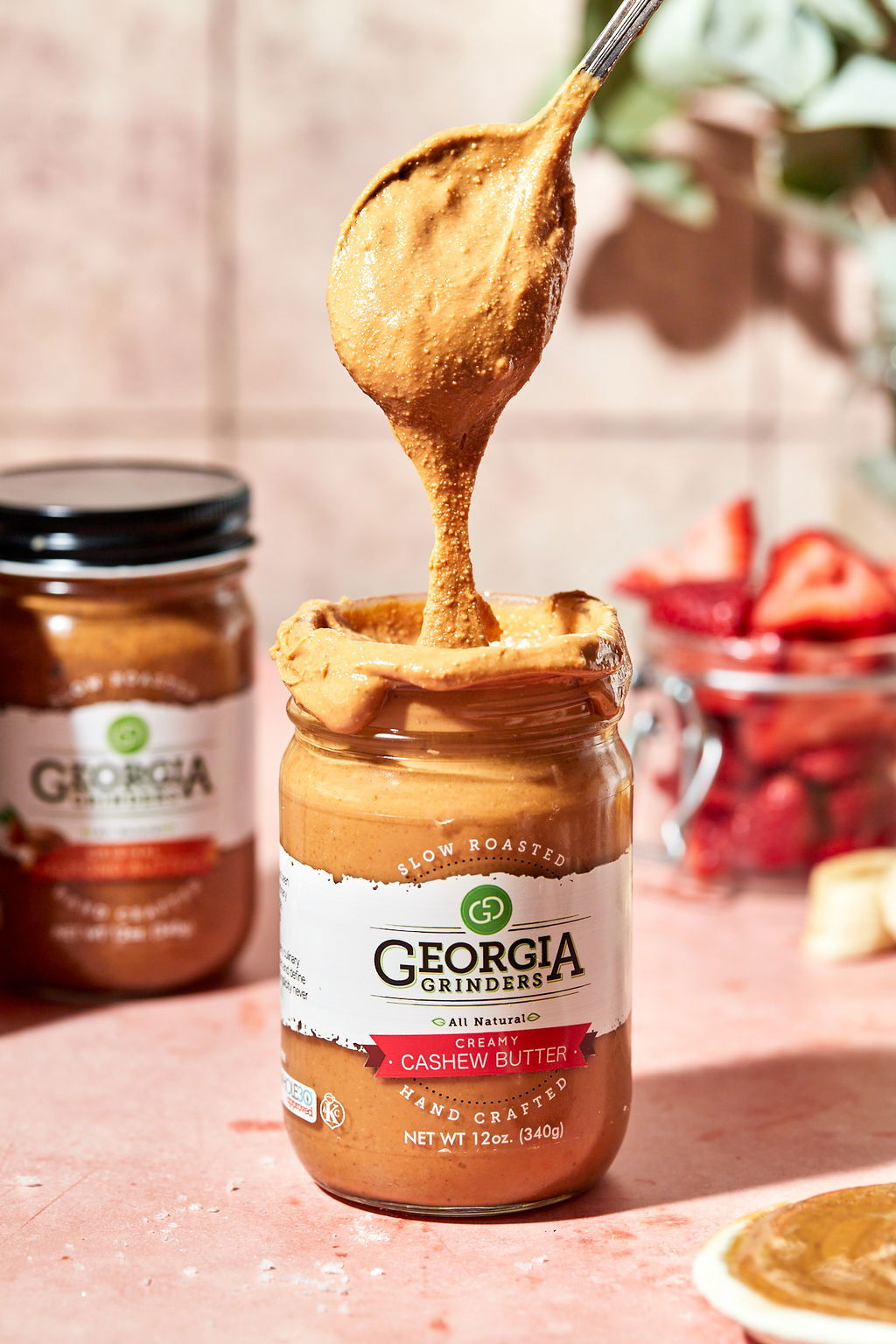 A 64 oz bulk tub of Georgia Grinders Cashew Butter (CP-CL) by Georgia Grinders, showing a spoon scooping out creamy, thick butter. Sliced strawberries and another tub appear in the blurred background on a pink surface. Keto & vegan friendly.