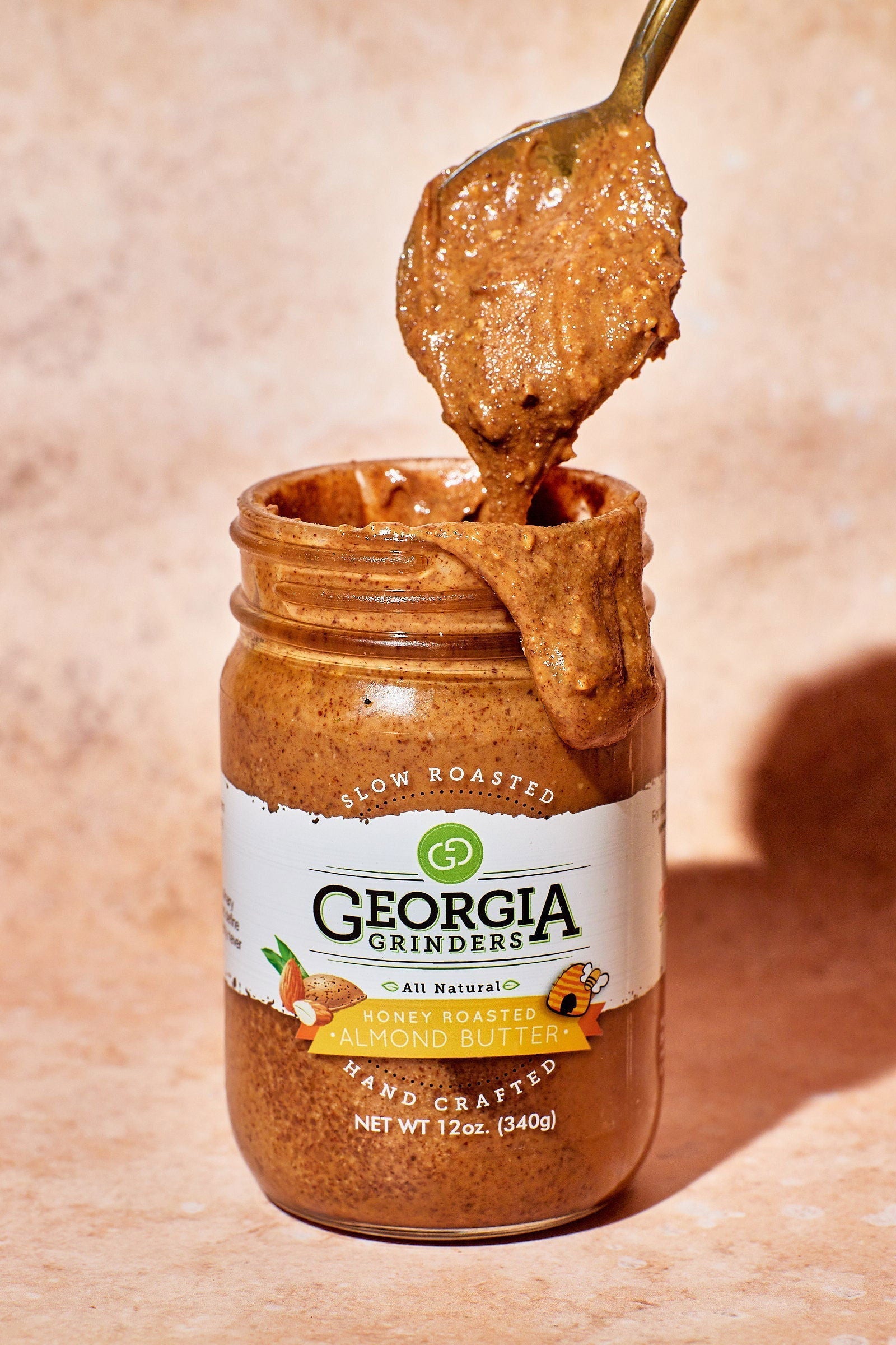 A spoon scoops thick, creamy almond butter from an open 64 oz bulk tub of Georgia Grinders Honey Roasted Almond Butter (CP-CL), a keto friendly treat, against a light beige background.