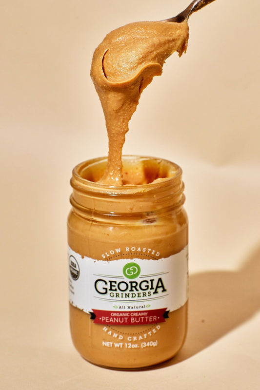 A spoon scoops keto-friendly Georgia Grinders Organic Creamy Peanut Butter from a nearly full 12 oz jar. The jar, part of the Georgia Grinders Organic Creamy Peanut Butter 4 Pack (CP-CL), is shown against a beige background.