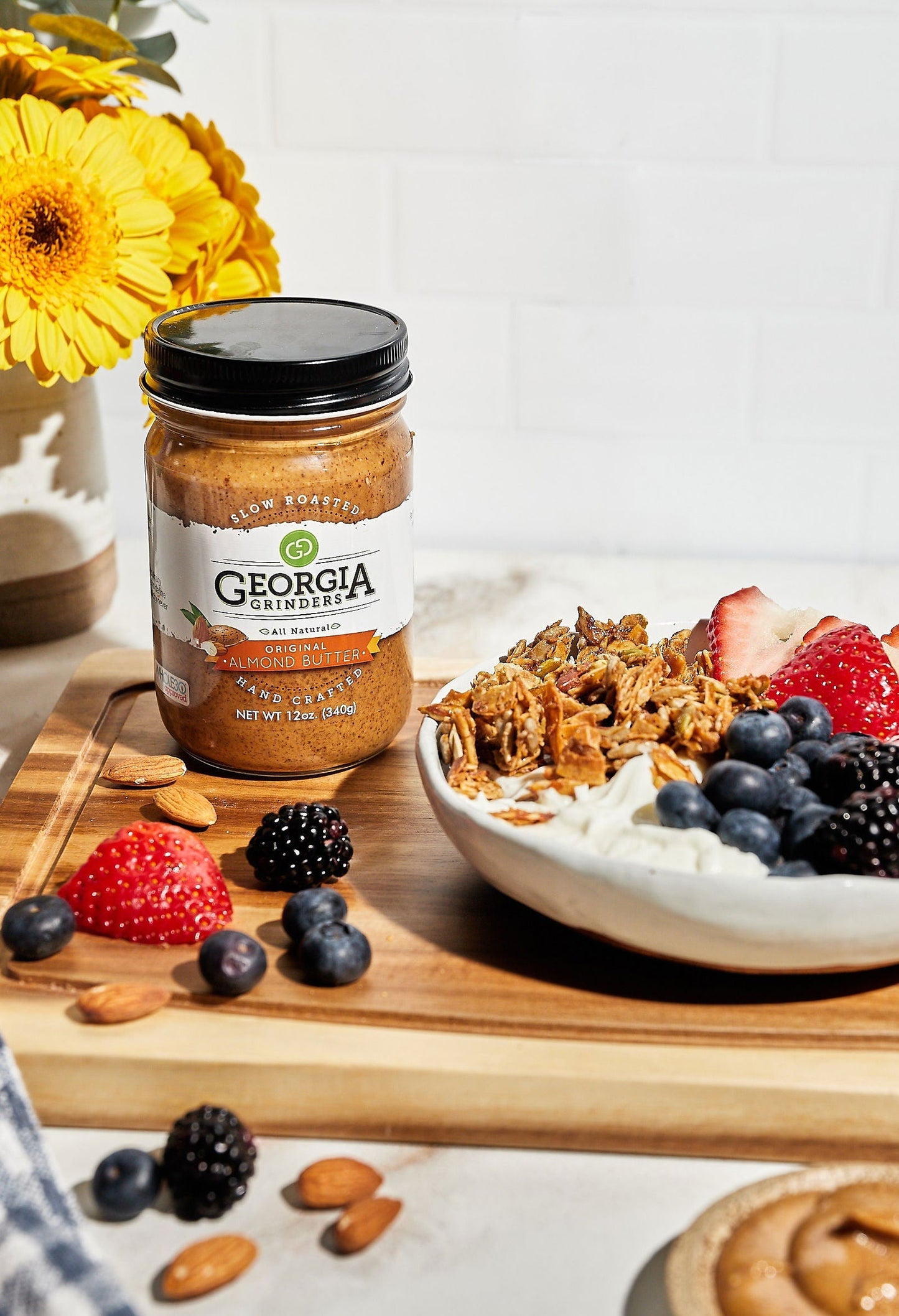 A 4-pack of Georgia Grinders Original Almond Butter (12 oz jars), by Georgia Grinders, sits on a wooden board beside granola, yogurt, strawberries, blueberries, and blackberries. Bright yellow flowers are in a vase behind them.