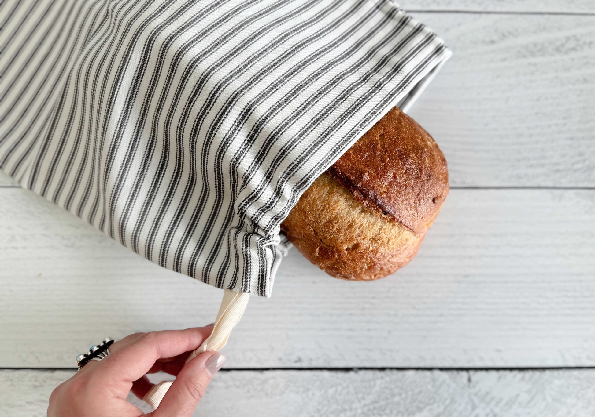A hand pulls the drawstring of a Dot and Army Grey Ticking Stripe Bread Bag, part of a set of two, holding a round homemade loaf on a white wooden surface.