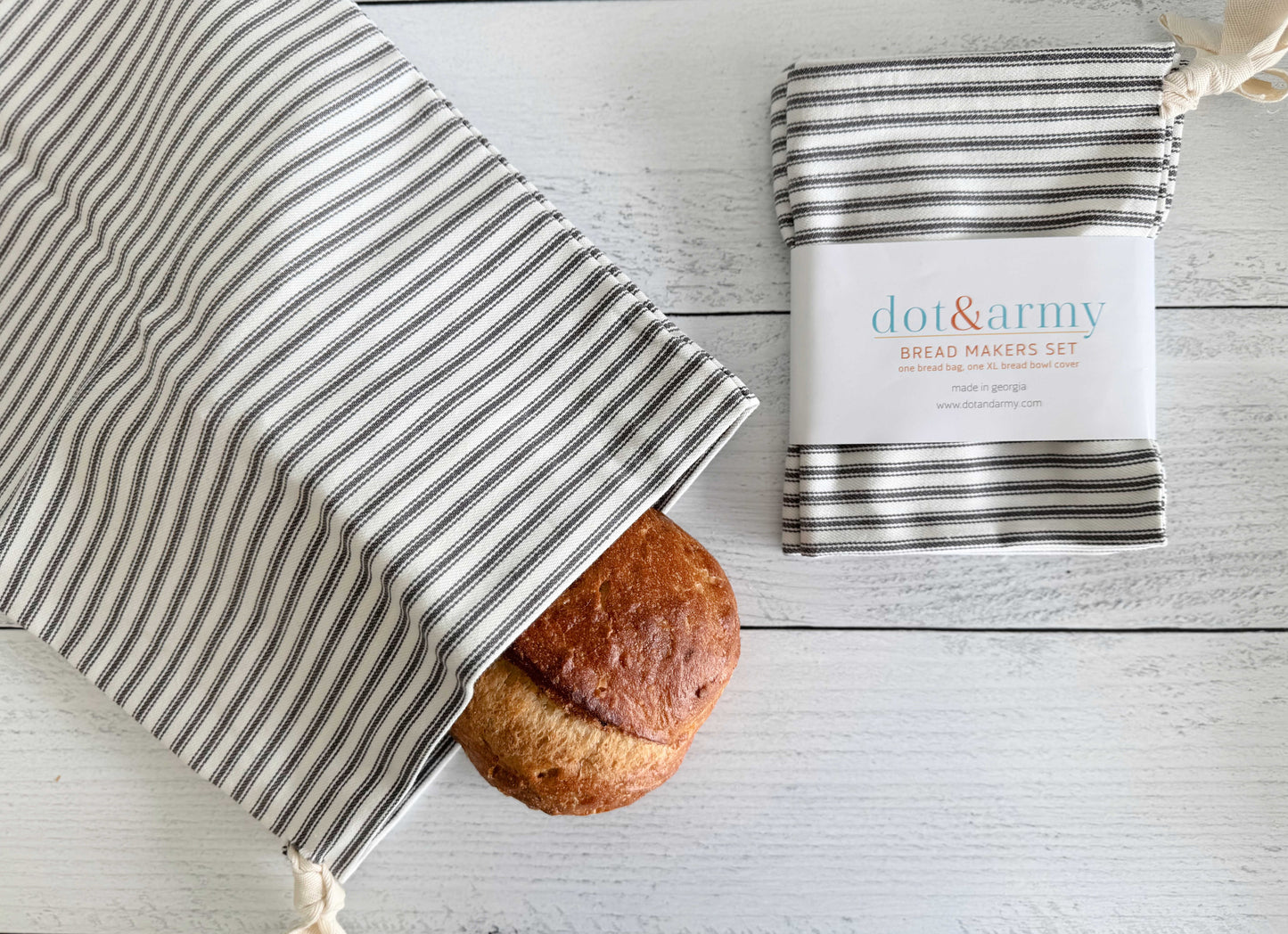 A round loaf of homemade bread peeks out of a Dot and Army Grey Ticking Stripe Bread Bag, with a matching folded cloth from the set of two beside it, all displayed on a white wooden surface.