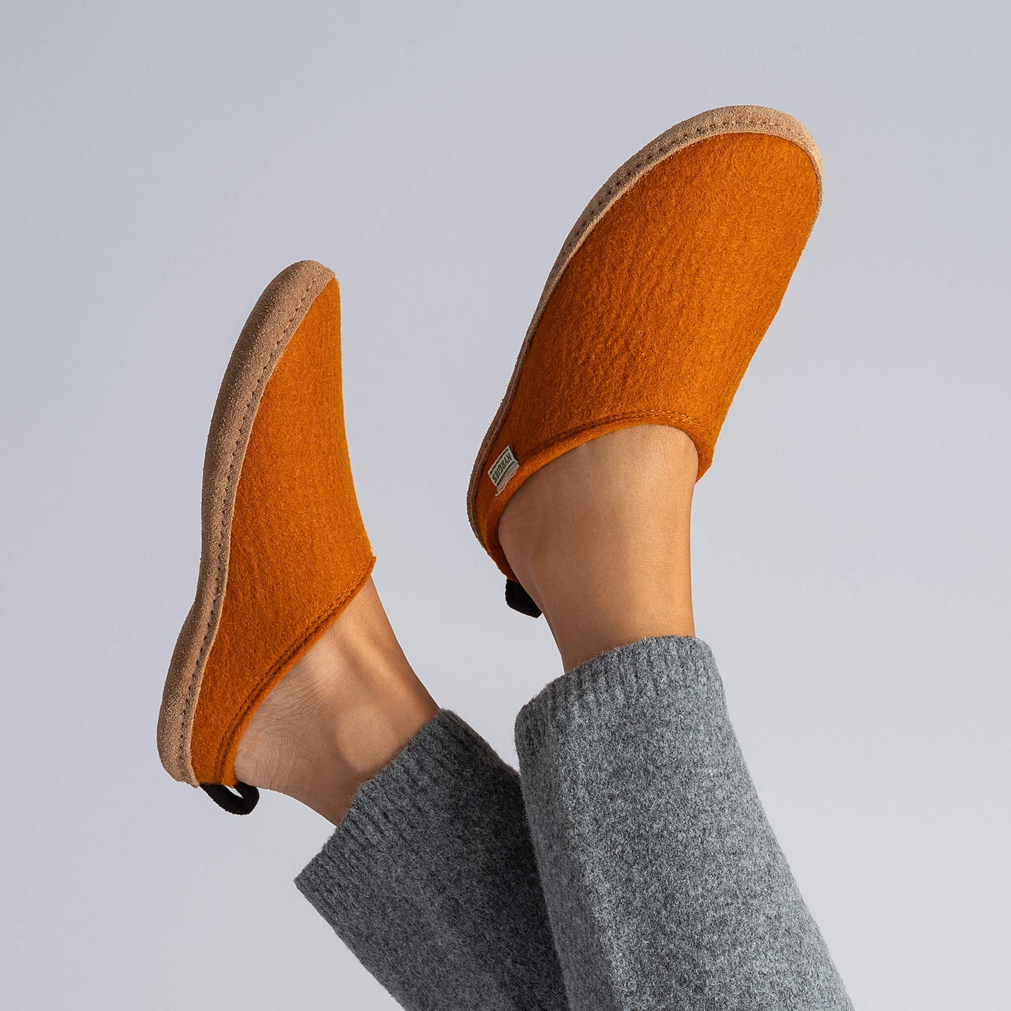 A person in grey knit pants lifts their feet, displaying orange Kyrgies Indoor Wool Slides by Kyrgies—handcrafted wool slippers with beige soles—set against a plain light background.