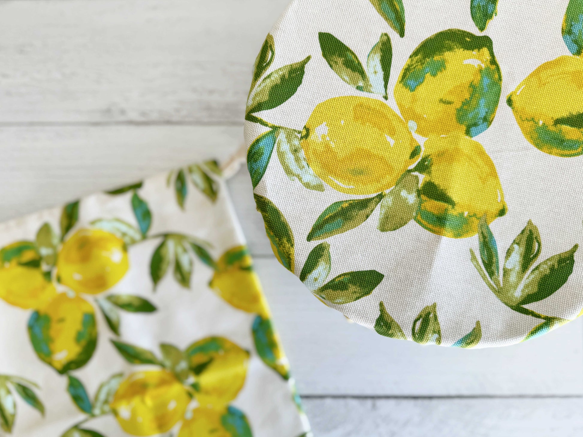 The Dot and Army Limoncello Bread Makers Set includes a bread bag and cover in white cotton, both featuring a bright yellow lemon and green leaf print—one covers a round container, while the other lies flat on a white wooden surface.