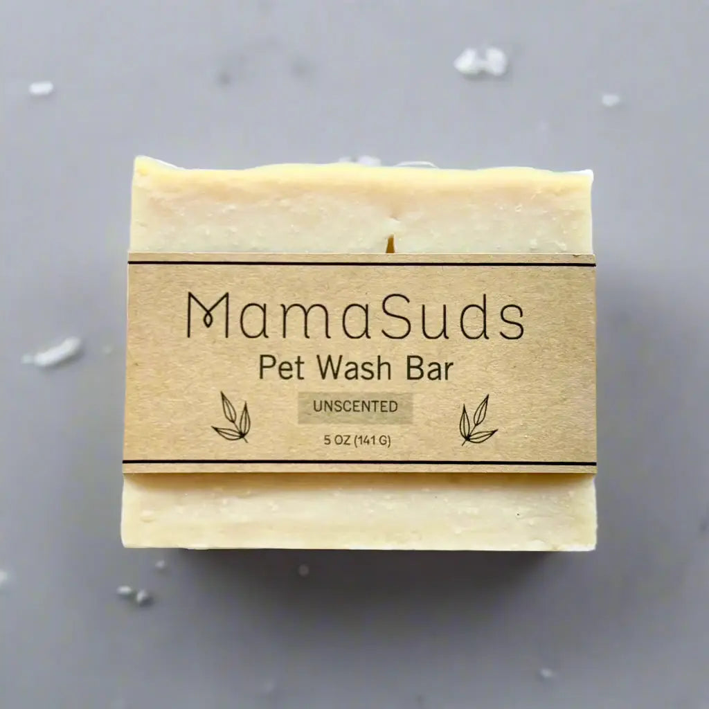 A 5 oz (141g) MamaSuds Pet Wash Bar Unscented rests on a gray surface with white soap flakes around it. This vegan, biodegradable soap is ideal for pets.
