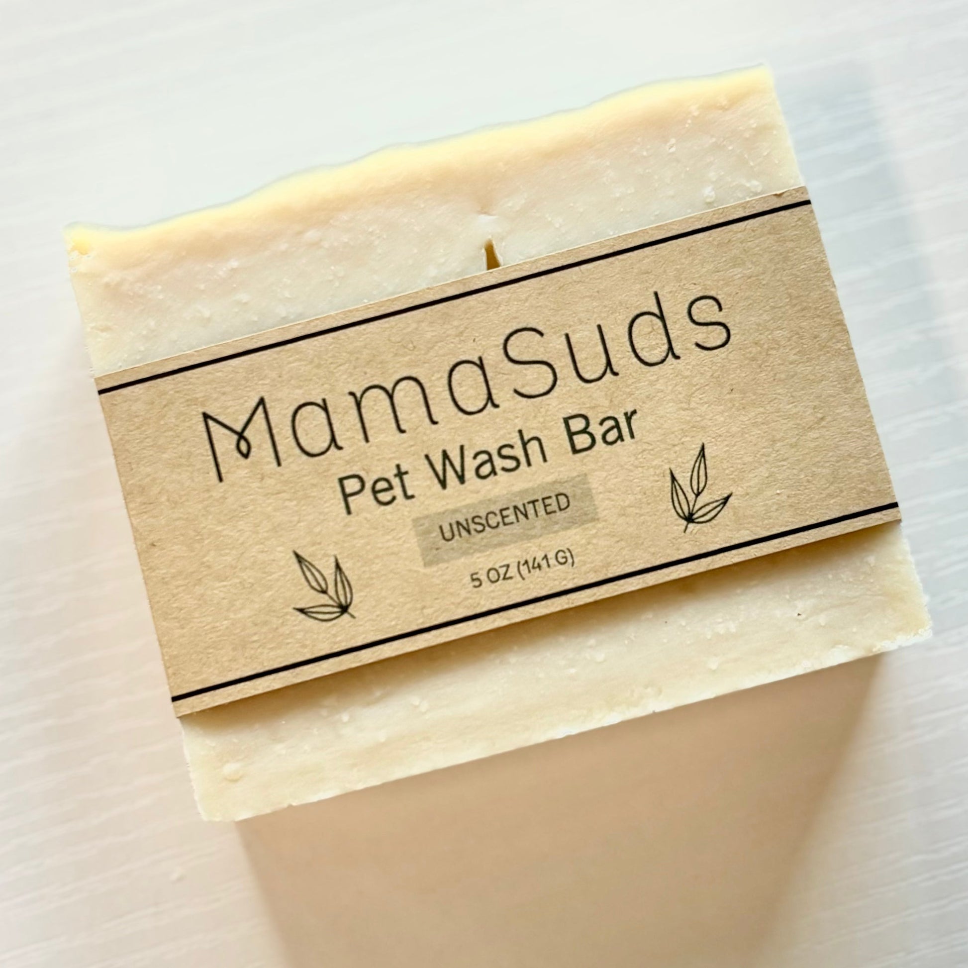 A beige, biodegradable vegan soap bar labeled MamaSuds Pet Wash Bar Unscented, 5 oz (141g) rests on a light textured surface.