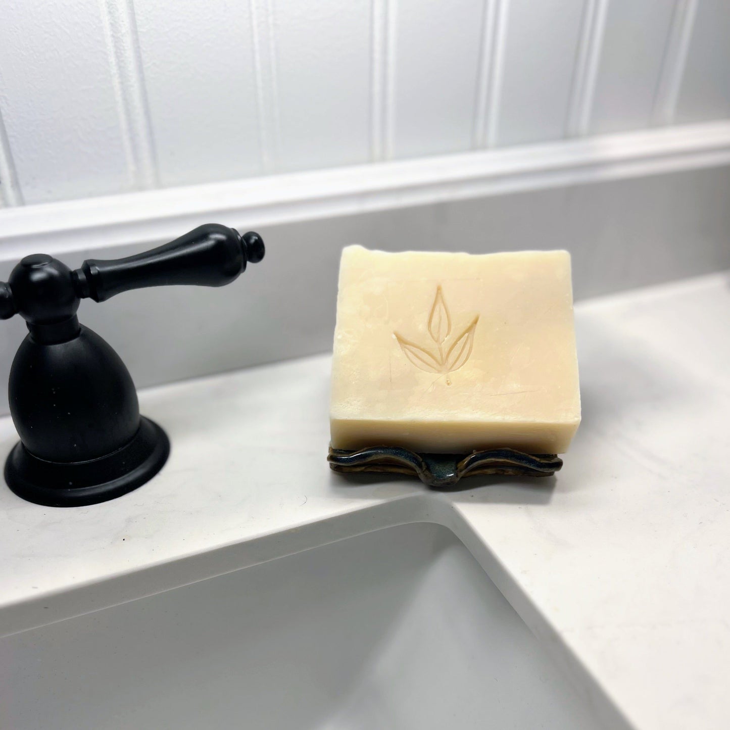 A bar of MamaSuds Scented Castile Bar Soap with a leaf design rests on a soap dish beside a black faucet atop a white bathroom sink with a paneled backsplash.