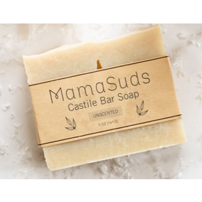 A creamy white Unscented Castile Bar Soap by MamaSuds, gentle for sensitive skin and biodegradable, rests on a light textured surface. It’s wrapped in a brown paper label featuring product details and simple leaf illustrations.