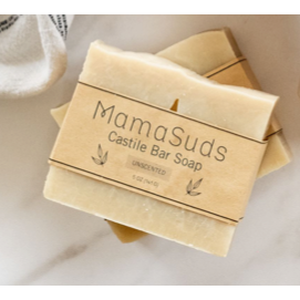 Two beige bars of MamaSuds Unscented Castile Bar Soap, perfect for sensitive skin, are stacked on a white surface with a towel behind them. This gentle, biodegradable soap cleanses without irritation.