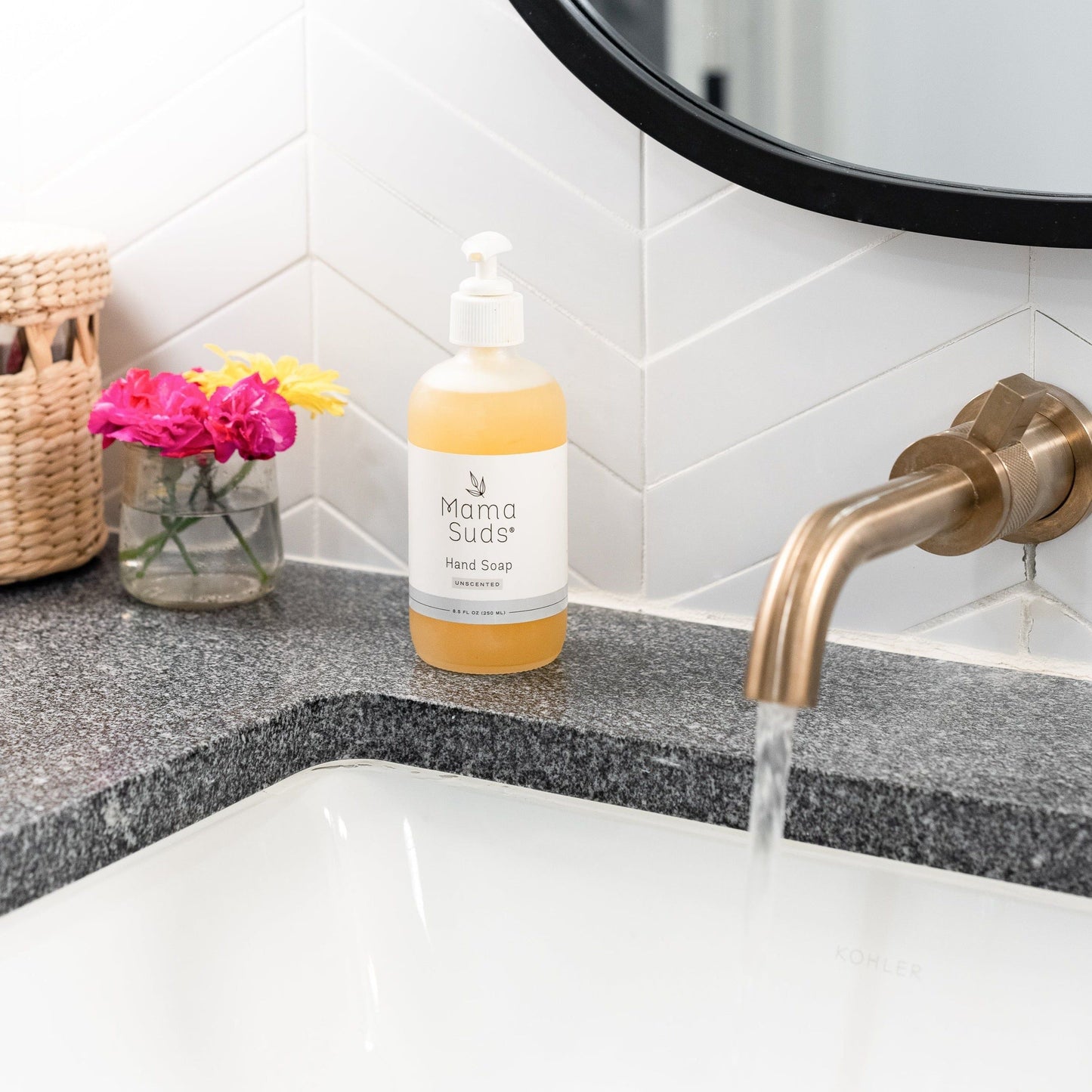 A bottle of MamaSuds Hand Soap sits on a gray countertop near a sink with running water, colorful flowers in a vase, and a woven basket, beneath a modern bathroom mirror and gold faucet.