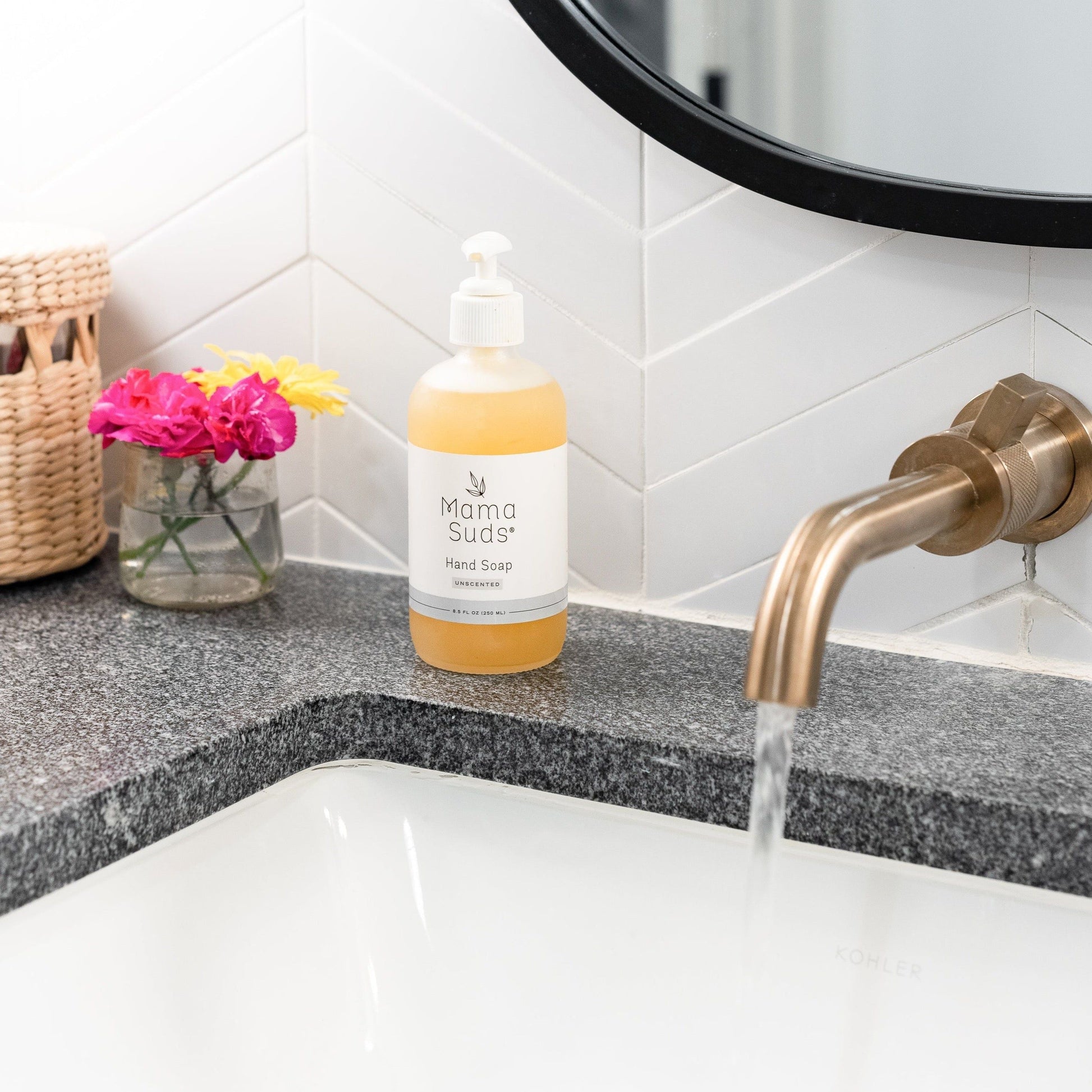 A bottle of MamaSuds Hand Soap sits on a gray countertop near a sink with running water, colorful flowers in a vase, and a woven basket, beneath a modern bathroom mirror and gold faucet.