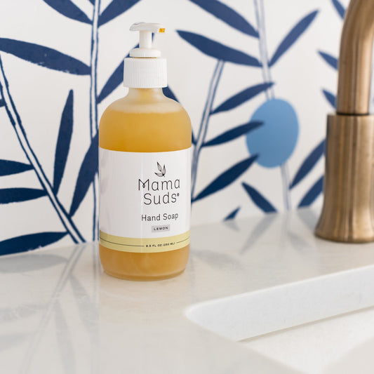 A bottle of MamaSuds Hand Soap with a pump dispenser sits on a white bathroom countertop beside a sink, set against a blue and white leafy patterned wall.