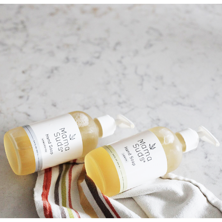 Two MamaSuds Hand Soap pump bottles rest on a light marble surface beside a colorful striped beige cloth. The biodegradable soap is yellow, and each bottle features a white label and pump.