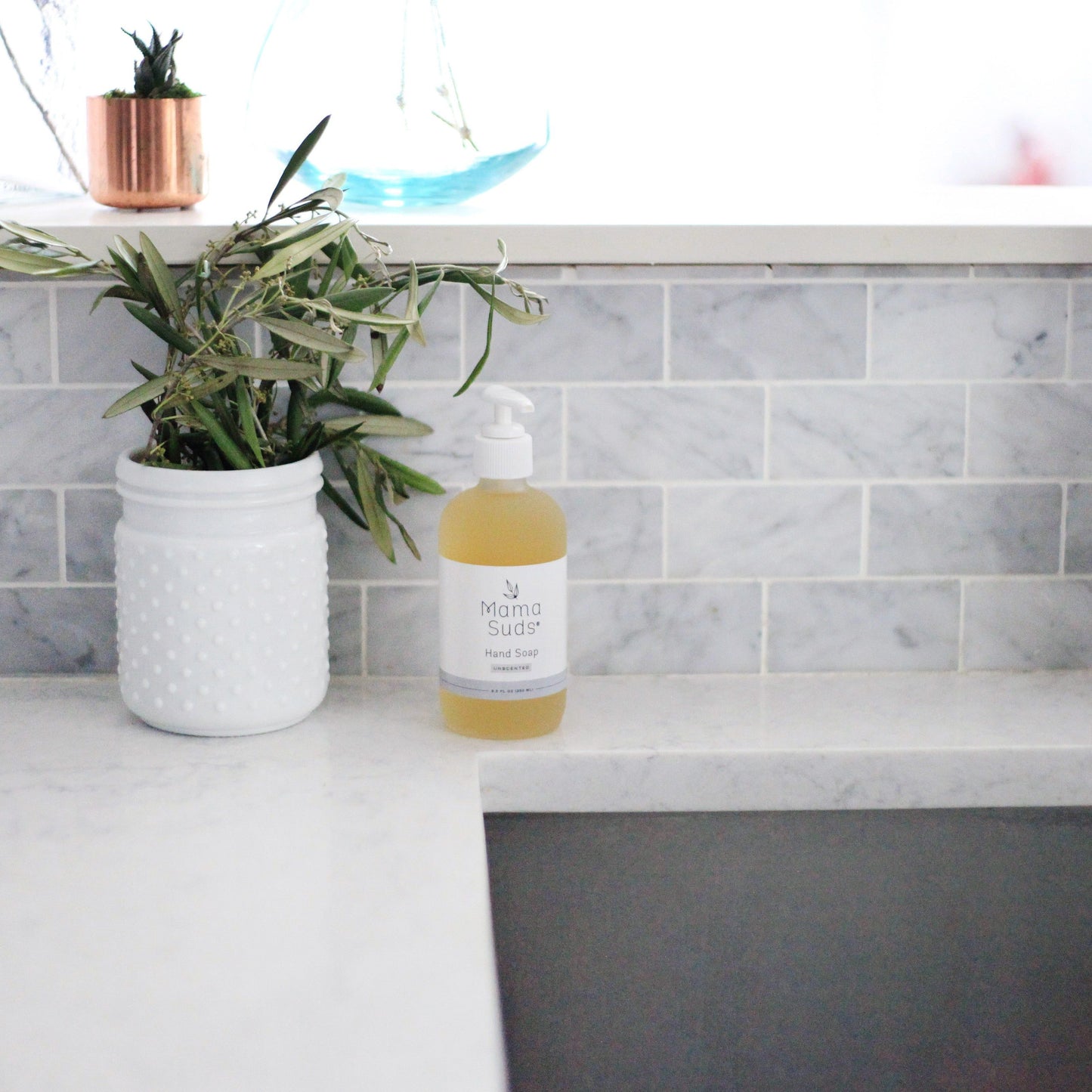 A bottle of MamaSuds Hand Soap sits on a white marble countertop beside a textured white jar with green foliage, set against a gray subway tile backsplash.