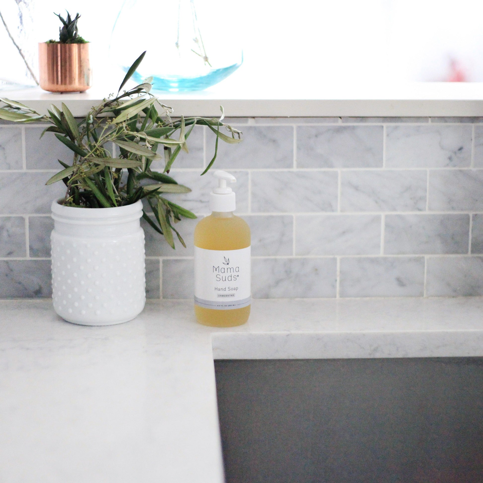 A bottle of MamaSuds Hand Soap sits on a white marble countertop beside a textured white jar with green foliage, set against a gray subway tile backsplash.
