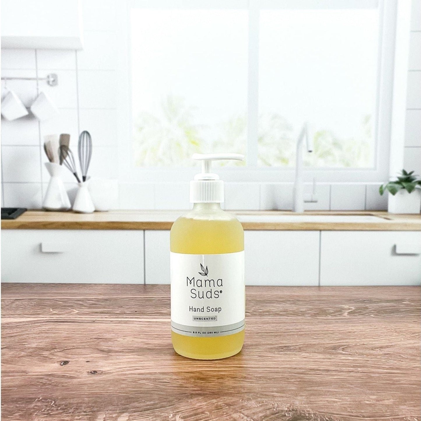 A bottle of MamaSuds Hand Soap rests on a wooden countertop in a bright, modern kitchen with white cabinets and a large window in the background.