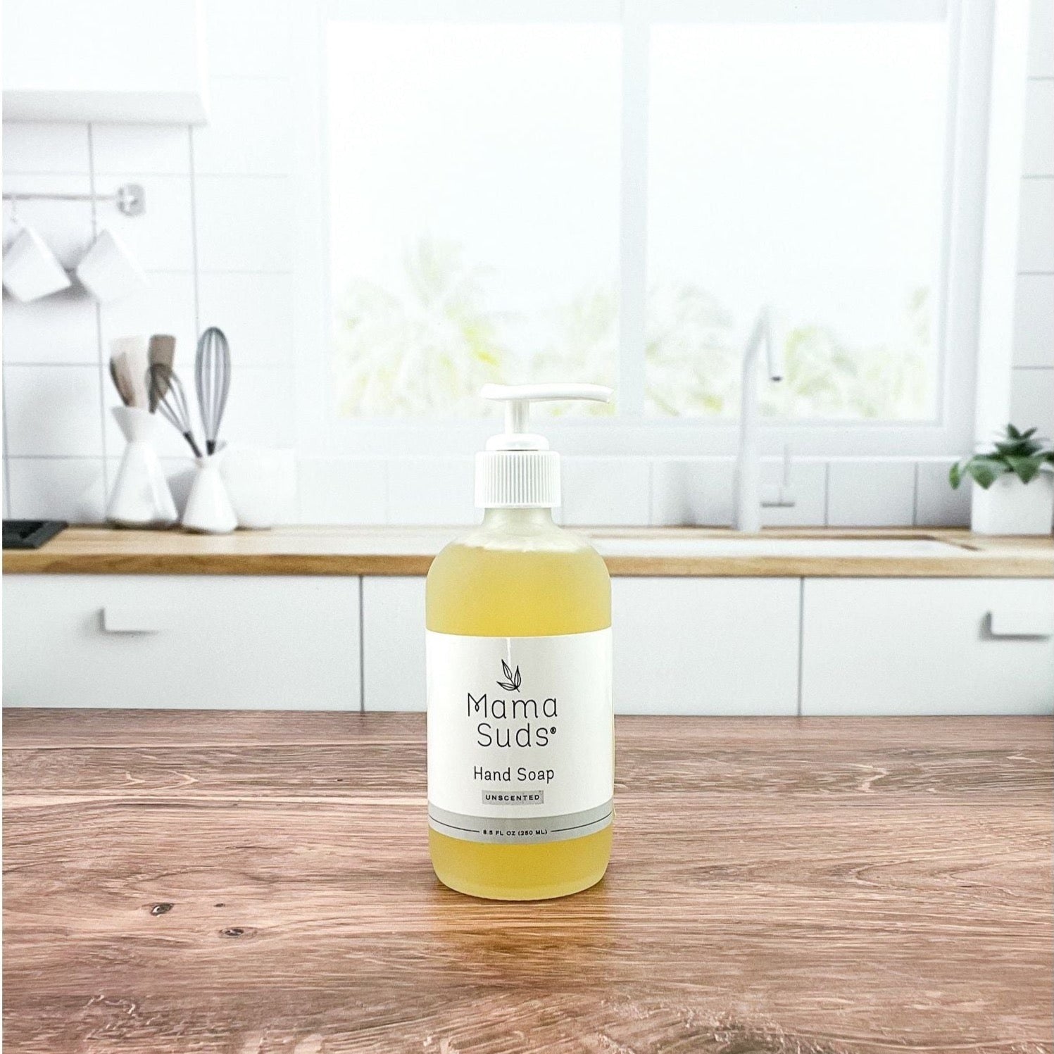 A bottle of MamaSuds Hand Soap rests on a wooden countertop in a bright, modern kitchen with white cabinets and a large window in the background.