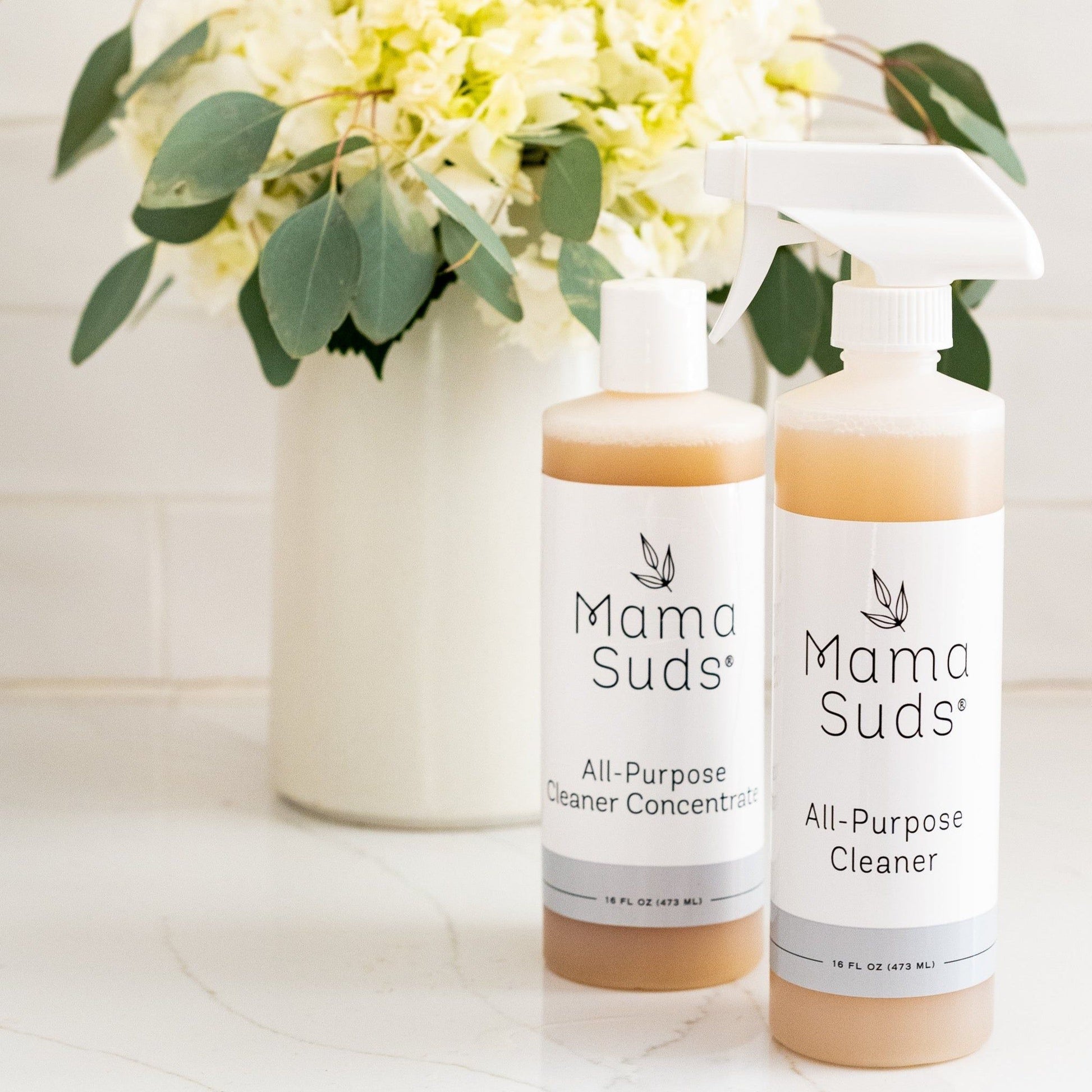 Two bottles of MamaSuds All-Purpose Cleaner CONCENTRATE, an eco-friendly cleaner, are on a white surface beside a white vase with light yellow flowers and green leaves.