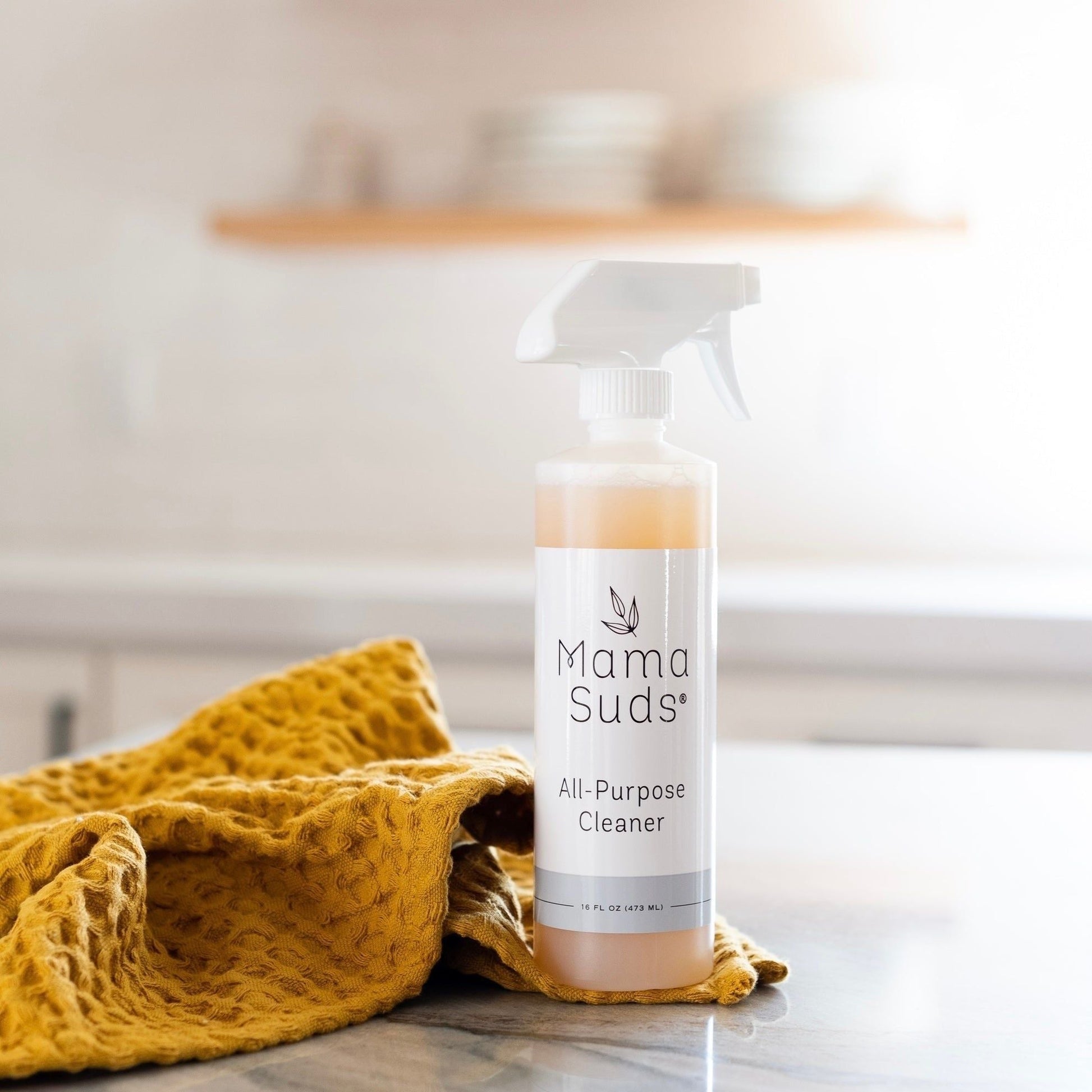 A bottle of MamaSuds All-Purpose Cleaner Spray, made with natural ingredients, rests on a countertop beside a textured yellow cloth, with blurred shelves and dishes in the background.