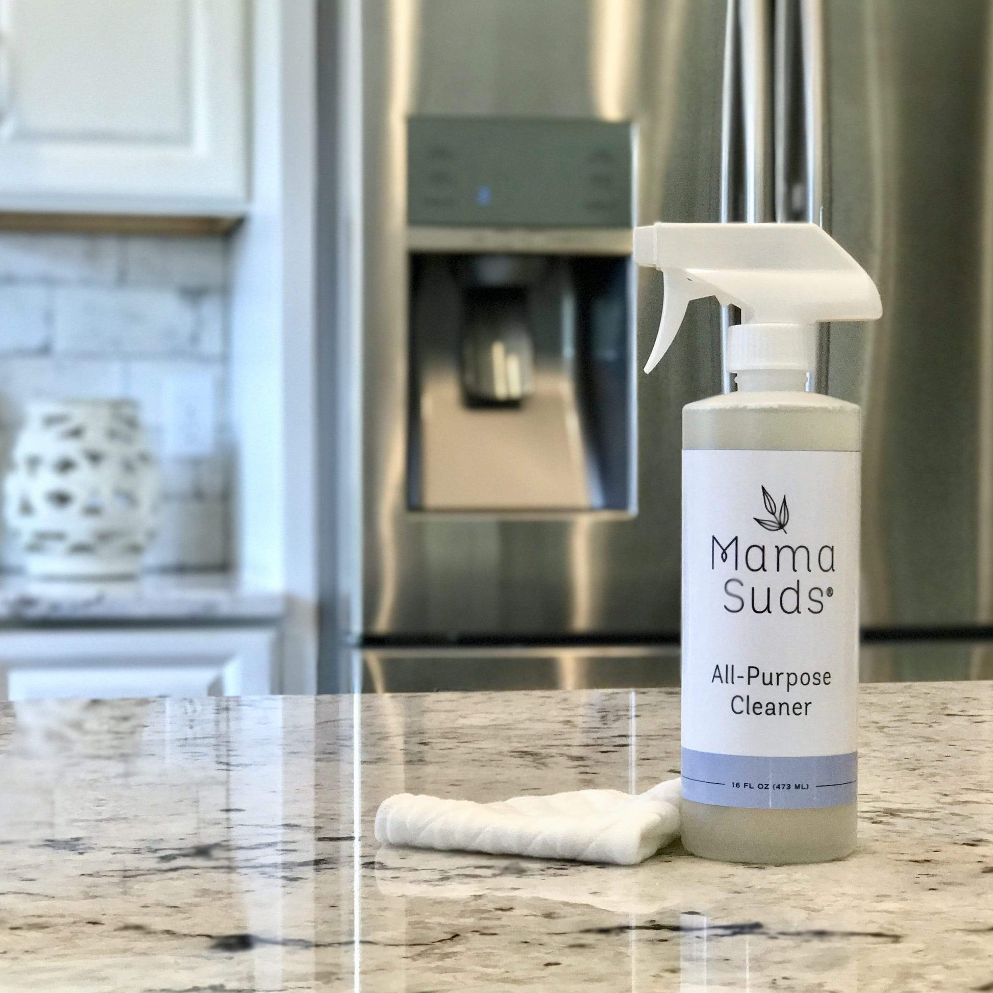A bottle of MamaSuds All-Purpose Cleaner Spray and a folded white cloth rest on a marble kitchen countertop, with a stainless steel refrigerator and white cabinets in the background.