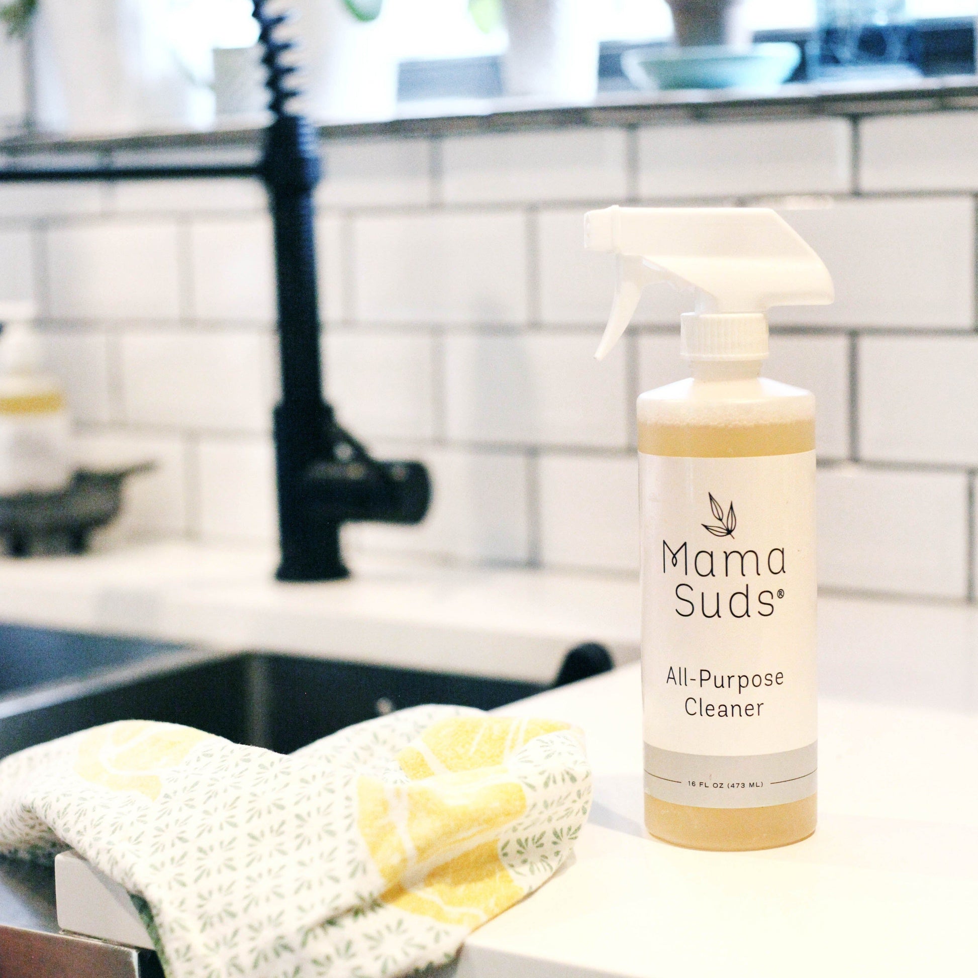 A bottle of MamaSuds All-Purpose Cleaner Spray, featuring natural ingredients, sits on a white kitchen countertop beside a patterned cloth, with a sink and tiled backsplash in the background.