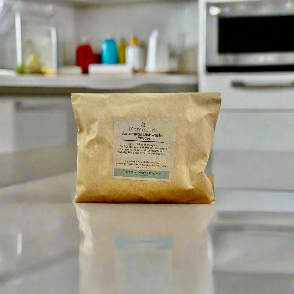 A brown paper bag labeled MamaSuds Automatic Dishwasher Powder SAMPLE, a biodegradable and cruelty-free cleaner, sits on a shiny kitchen counter with colorful items and an oven in the background.