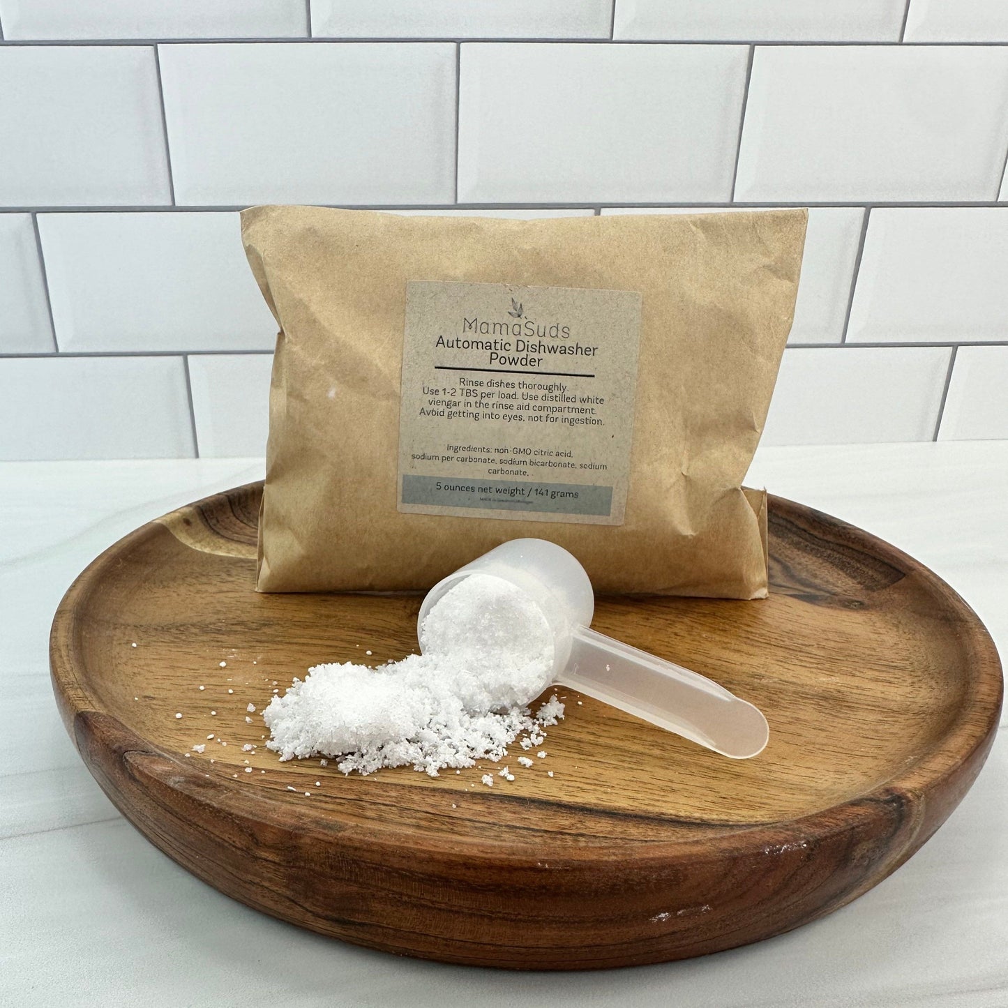 A brown paper bag labeled MamaSuds Automatic Dishwasher Powder SAMPLE, a cruelty-free and biodegradable product, sits on a wooden tray with a plastic scoop of white powder in front, set against white tiles.