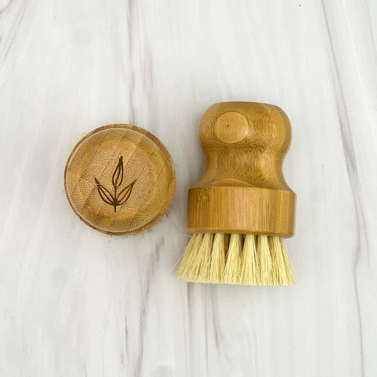 The MamaSuds Bamboo Pot Brush with a wooden handle and natural sisal bristles is shown beside its matching sustainable bamboo lid, which features an engraved leaf design. Both rest on a light surface.
